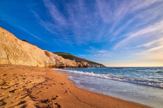 Beautiful low sun view of secluded and remote beach on sunny evening. High yellow cliffs, weathered rocks, azure sea waters, sandy beach, clear sky, no people. Agios Ioannis beach and Aegean sea, Milos island, Greece.