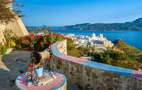 Colorful view on traditional Greek town by harbour from decorated terrace with well, bucket, rich red cranesbills in evening. Whitewashed houses, Orthodox Greek church, blue sea water, far mountains, clear sky, low sun, sunset, Plaka, Milos, Greece