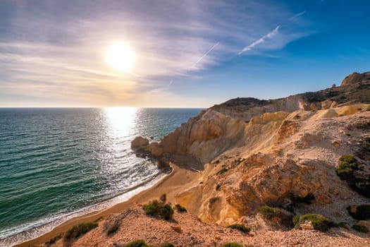 Beautiful low sun view of secluded and remote beach on sunny evening. High yellow cliffs, weathered rocks, azure sea waters, sandy beach, clear sky, no people. Agios Ioannis beach and Aegean sea, Milos island, Greece.