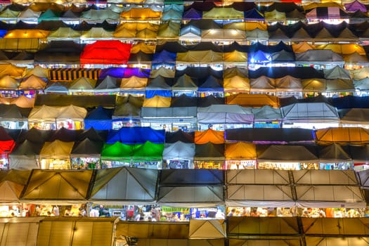Close-up view of rows of colorful outdoor market tents and food stalls at night. Fairs, marketplaces, small businesses, shopping, dining, Asian markets, local traditions, tourist attractions. Ratchada Rot Fai train night market, Bangkok, Thailand.