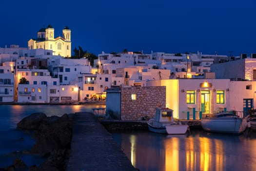 Traditional Greek whitewashed houses and church of Dormition of Mother of God on hilltop by seafront after sunset, Naoussa, Paros island, Greece. Colorful water reflections, pier and boats moored in harbour.