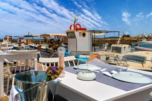 Tables and chairs of seafood restaurant by waterfront of Naoussa bay, fishing boats moored next to tables in summer day sunshine. Traditional Greek cuisine, vacations, travel destination, tourist paradise, romantic dining, selective focus.