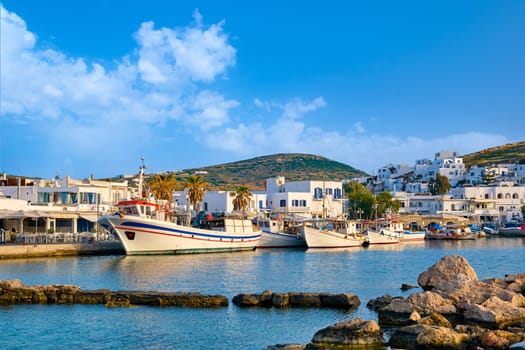 Beautiful tradition Greek fishermen village on hilltop. Colorful boats moored by jetty, whitewashed houses, evening or morning sunlight, clear blue sky, blue sea waters. Travel destination, vacation paradise, summer vibe.