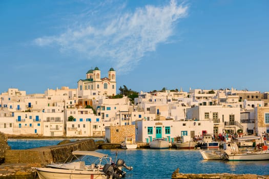 Traditional Greek whitewashed houses and church of Dormition of Mother of God as local landmark on hilltop by seafront on sunny day, Naoussa, Paros island, Greece. Pier and boats moored in harbour, clear blue sky, summer sunshine, cafes and bars.