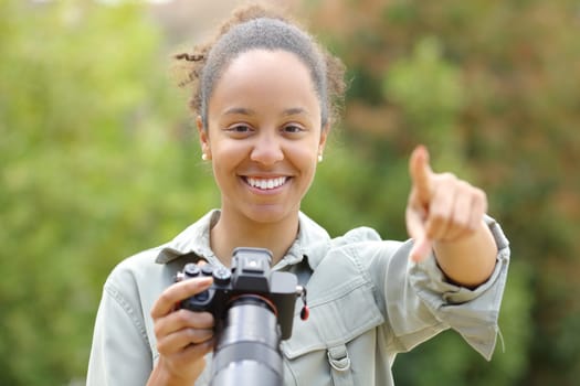 Front view portrait of a black photographer pointing at camera holding mirrorless camera in a park
