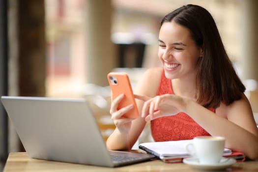 Happy student checking smart phone and computer in a bar terrace
