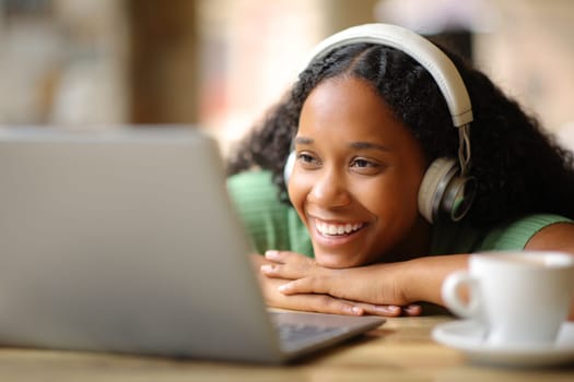 Happy black woman wearing headphone watching media on laptop in a bar terrace
