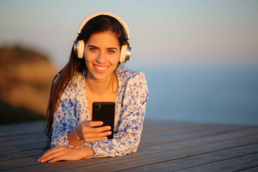 Front view portrait of a happy woman wearing headphone listening to music on the beach looking at you