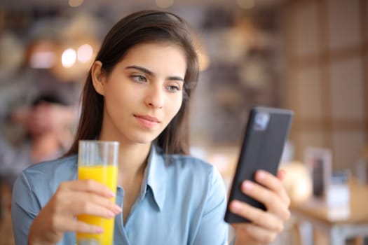 Woman drinking orange juice using phone sitting in a bar at breakfast