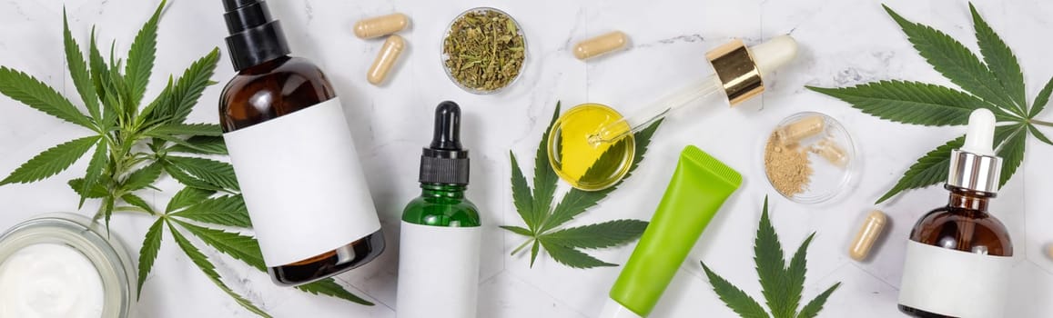 Cosmetic bottles with blank labes, Pipette with CBD oil, hemp capsules and green cannabis sativa leaves on a marble table top view, Mockup, Copy space. Organic skincare beauty products.