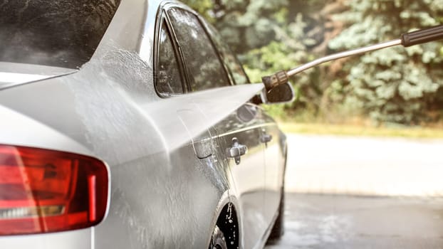 Side of silver car washed in self serve carwash, jet water stream spraying, sun lit trees background