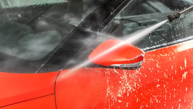 Side mirror of red car washed in self serve carwash, jet water stream spraying from hose nozzle.