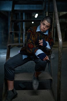 Young woman sitting on steel ladder with smoldering cigarette and bottle of beer. Dregs of society concept