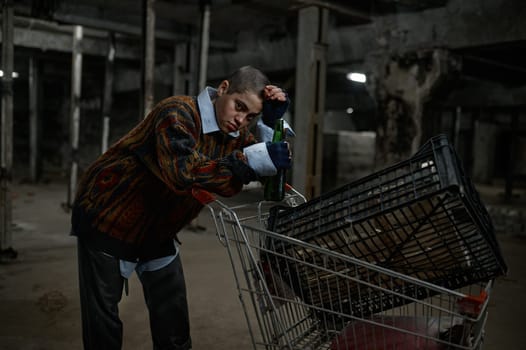 Portrait of unhappy young woman immigrant or beggar drinking beer leaned on trolley cart with her belongings