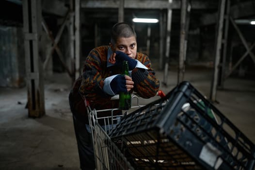 Portrait of unhappy young woman immigrant or beggar drinking beer leaned on trolley cart with her belongings