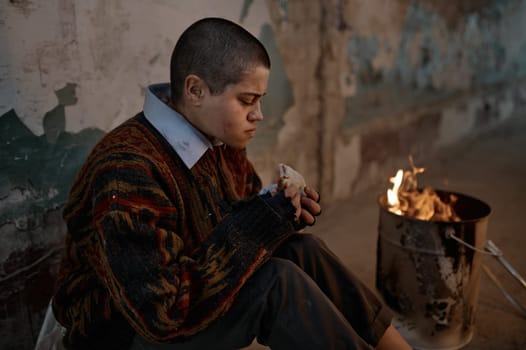 Poor homeless hungry woman eating sandwich while warming at bonfire burning flame