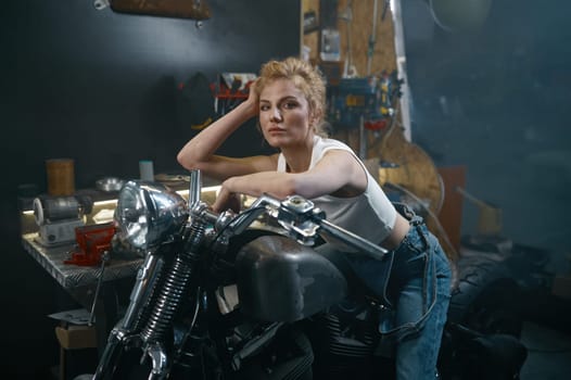 Portrait of beautiful woman biker sitting on motorcycle in garage. Beautiful female mechanic posing for camera at workshop
