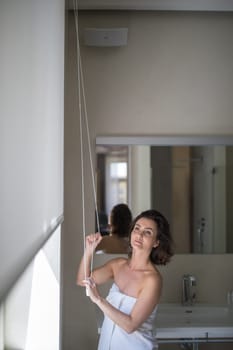 Middle-aged woman opening white roller blinds on window in a modern apartement