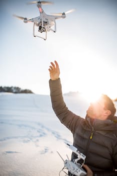 Young man controlling his drone in snowy outdoors. Drone operator holding a transmitter and landing with a drone.