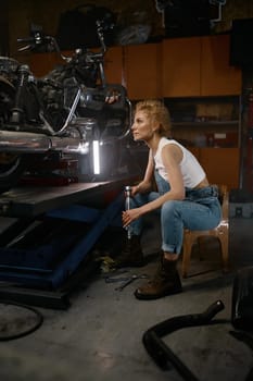 Thoughtful young woman holding ratchet spanner brainstorming trying to solve problem with custom motorcycle. Workshop interior in evening