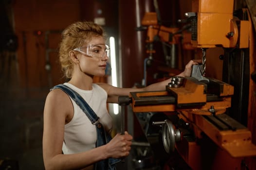 Close up view of focused professional serious young woman wearing safety protective goggles working on electric milling and drilling machine at garage workshop