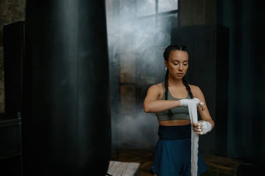 Closeup atmospheric shot of woman boxer wearing tracksuit wrapping her hands in bandage standing nearby punching bag at vintage old sport gym club