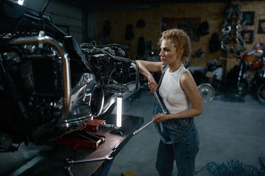 Talented girl wearing blue jumpsuit using work tool to repair motorcycle in garage. Pensive young woman at creative authentic workshop