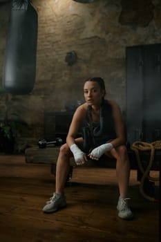 Young sportive female boxer in boxing bandage sitting on bench over loft gym interior. Kickboxing training club for woman