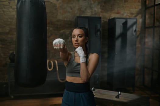 Front view portrait of powerful serious woman boxer in boxing bandage fighting to camera making direct hit. Young fit female fighter training punches workouting at gym