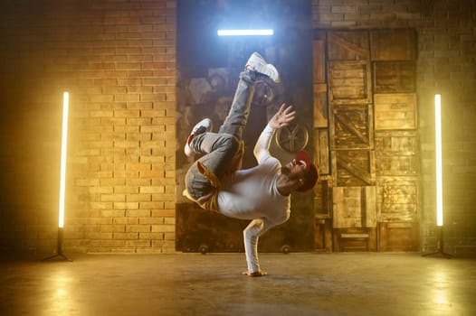 Young man breakdancing doing one handed handstand. Active stylish guy hip hop dancer training or performing over loft studio background