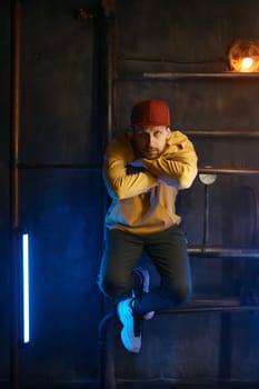 Hipster breakdancer wearing casual clothes posing for camera over loft underground subway background. Portrait of B-boy hip hop dancer