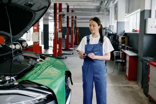 Young woman mechanic or car engineer wearing uniform overalls writing on clipboard at car service