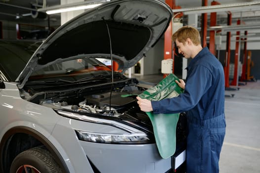 Male mechanic wearing blue uniform using protective rubber cape for work in engine repair service standing front of car opened hood