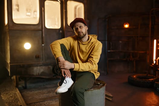 Young male breakdancer sitting on steel barrel tank in loft dance studio