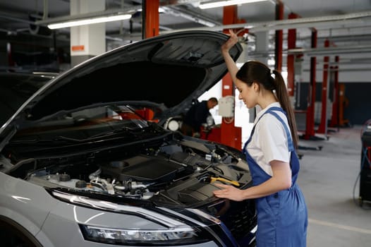 Young woman car mechanic looking under hood while working at repair garage. Female technician trying to solve problem of inoperative engine