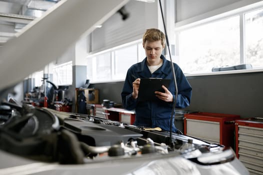Young male mechanic of service station wearing uniform standing in front of opened car hood holding clipboard in hand writing notes during automobile examination