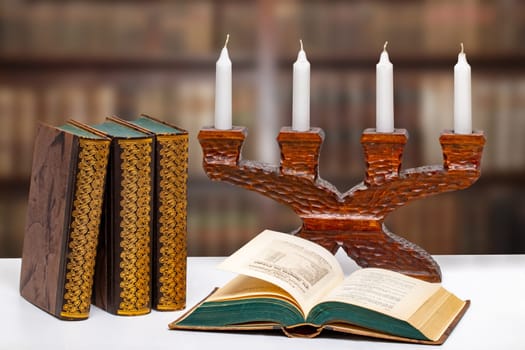 Antique books with an open book in front of an antique wooden candlestick on a bright table over abstract blurred library. University, history, memory and knowledge concept.