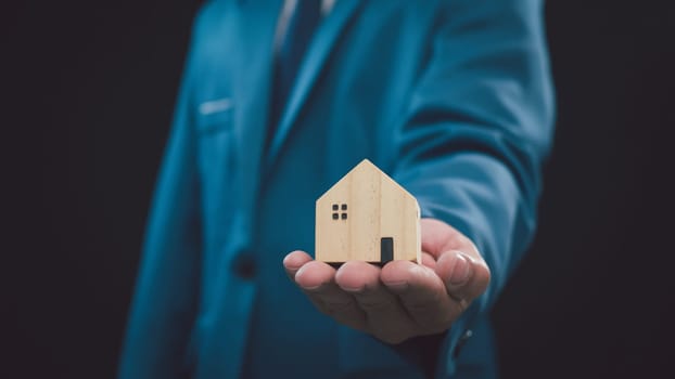 Concept of selling a house. Businessman is holding a model house on a dark background. Real estate agent offer house, property insurance and security, affordable housing concepts, home insurance broker agent, salesman person.