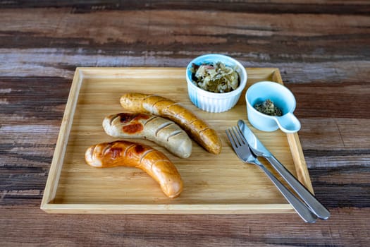 Photos of Grilled German Sausages with sauerkraut and dijon mustard in small bowls with knife and fork Together in a rectangular wooden tray, on a wood grain table. Ready to serve. Selective focus.