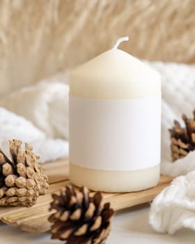 Candle with blank paper label on wooden tray near pine cones and sweater, Close up, copy space. Boho winter packaging mock up. Christmas Home decorations, Bohemian or scandinavian style
