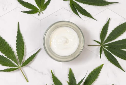 Cream jar near green cannabis sativa leaves on a marble table top view. CBD Cosmetic, Organic skincare beauty product. Eco friendly body or hand cream with hemp