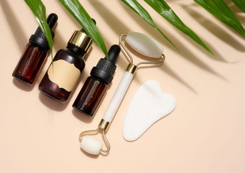 Brown glass bottle with a pipette, a white stone hand massager and a scraper for cosmetic procedures on a beige background, top view