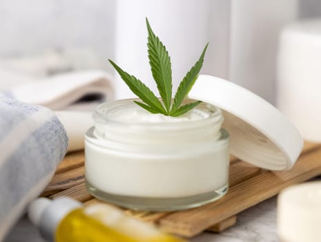 Opened cream jar with green cannabis leaves on tray close up in bathroom. Organic skincare beauty products, alternative CBD cosmetics
