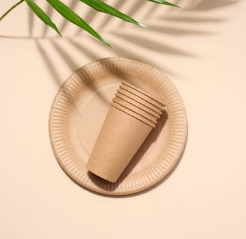 Brown paper plate, stack of paper cups, disposable tableware on a beige background, top view