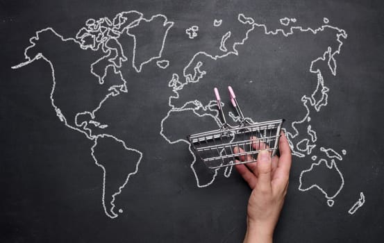 A female hand holding a miniature empty shopping basket against the background of a drawn world map. Concept of online shopping, international shipping