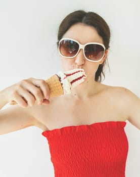 Happy cheerful girl in a red top and white sunglasses eats a colorful ice cream cone in her hands and smiles. Cinematic photo filter