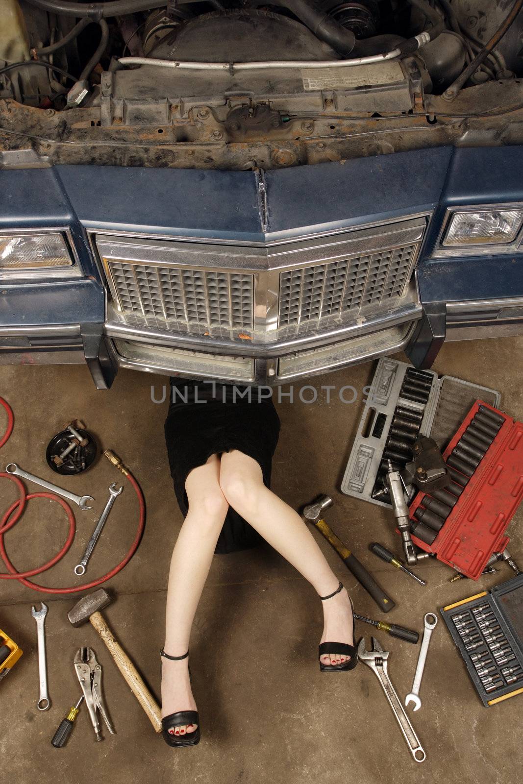 A female wearing a black skirt and heels doing repairs under the front of an old car from the early 80's.