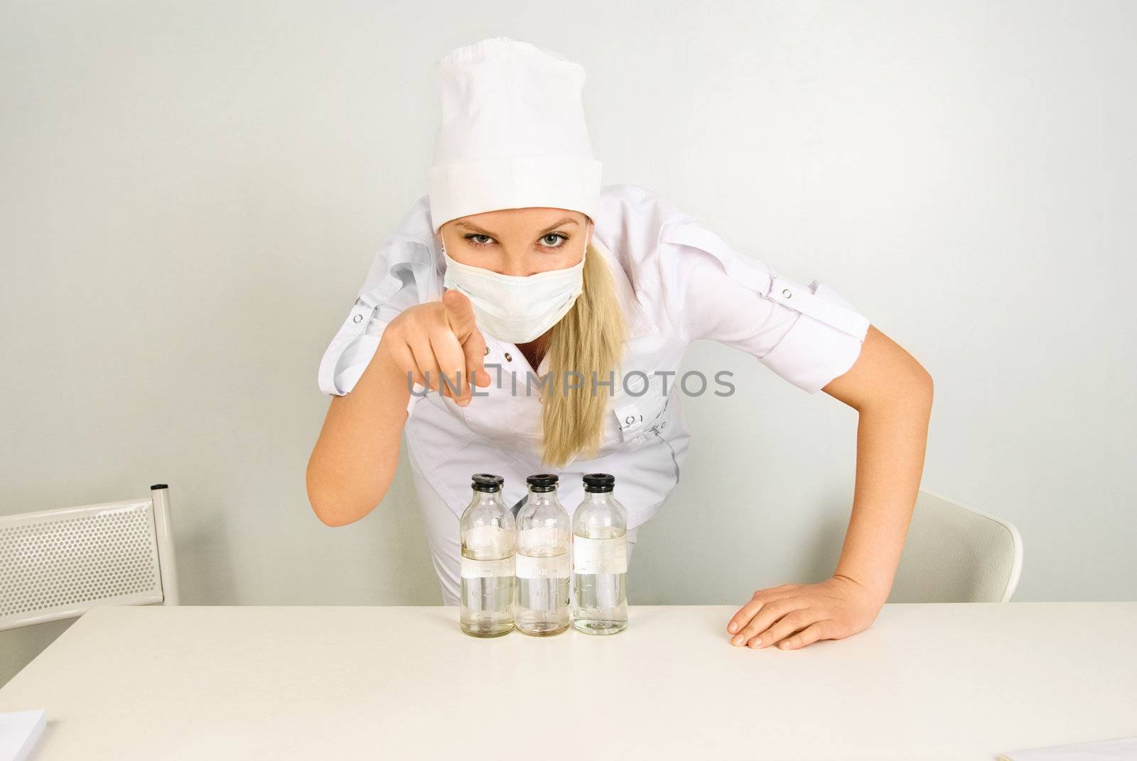 portrait of a young doctor with medications sitting by the table in her office and pointing at us with a finger
