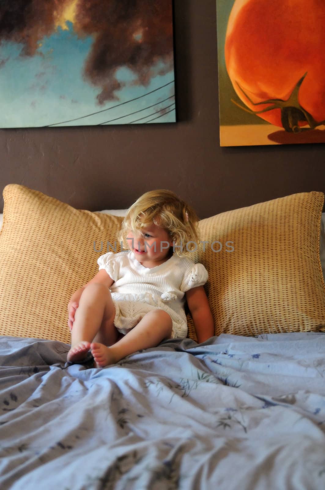 A blonde female toddler in a white dress sits on her parents' bed, grinning and drawing her knees up towards her chest.  The image is in selective focus.