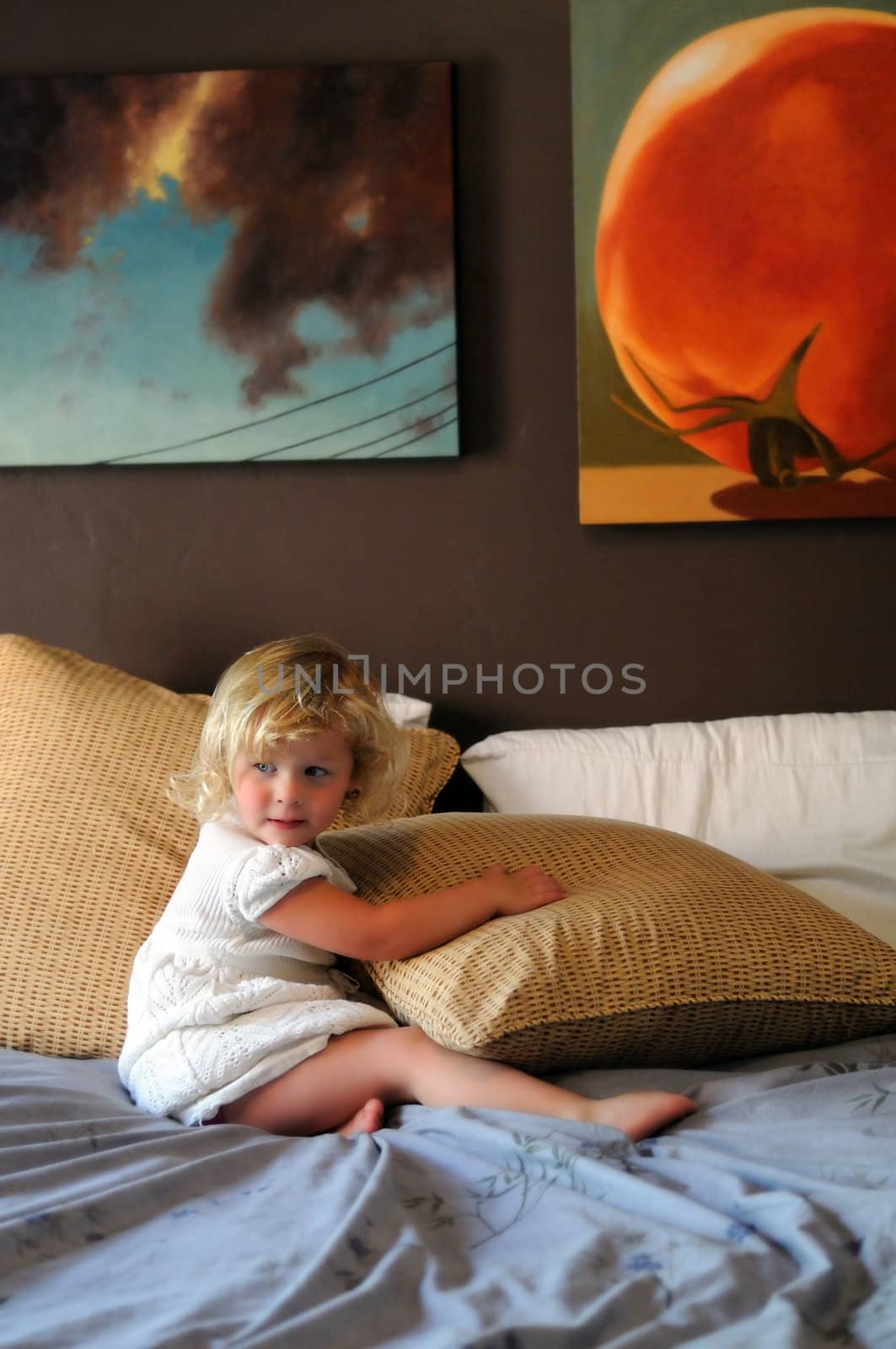 A blonde female toddler in a white dress sits on her parents' bed, turning away to her right.  The image is in selective focus.